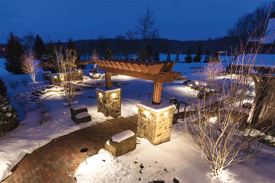 A snow-covered outdoor pathway with stone pillars and soft lighting, leading under a wooden pergola in a winter evening setting.