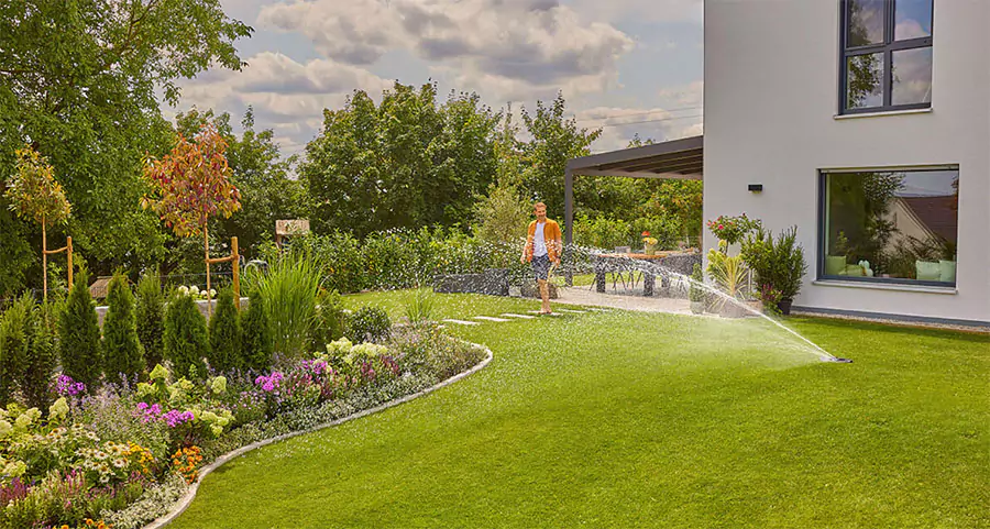 Homeowner inspects a sprinkler system watering a lush lawn and vibrant flower beds during a summer irrigation checkup in Indianapolis, IN.