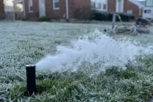 Sprinkler head spraying water over a frost-covered lawn in front of a brick home on a cold morning