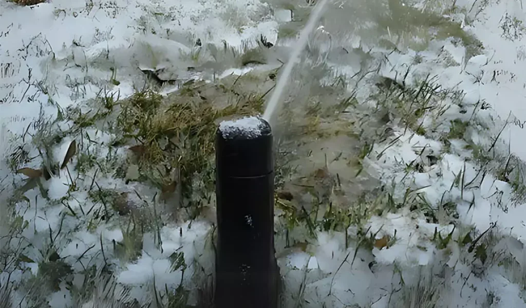 Irrigation system sprinkler head spraying water in a snow-covered lawn, highlighting winter preparation for irrigation systems.